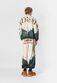 Conjunto de sudadera y pantalones en beige con estampado de paisaje verde. Presenta letras grandes, puños elásticos y bolsillos. Botas color camel completan el look.