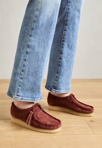 Mocassins en cuir suédé marron à bout rond, avec un design à lacets et un détail de pompon, associés à un jean bleu clair à ourlet effiloché.