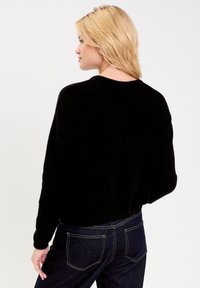 Maglione nero oversized con scollatura tonda e maniche lunghe, realizzato in morbido materiale, indossato con jeans blu scuro. Design minimalista.