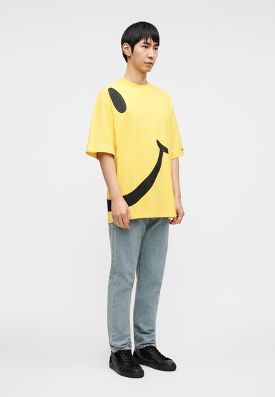 Κίτρινο βαμβακερό t-shirt με μεγάλο μαύρο γραφικό πρ όσωπο χαμόγελου, κοντά μανίκια, άνετη εφαρμογή. Συνδυάζεται με ανοιχτόχρωμα τζιν παντελόνια και μαύρα αθλητικά παπούτσια.