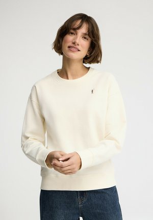 Vrouw met kort bruin haar, gekleed in een crèmekleurige trui met een klein geborduurd logo en een blauwe spijkerbroek, staand tegen een effen witte achtergrond.