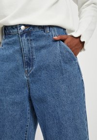 Jeansy z wysokim stanem w niebieskim kolorze, o prostym kroju, z przednimi kieszeniami, metalowym zapięciem na guzik oraz subtelnymi detalami szwów.