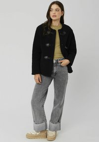 Veste extérieure noire texturée avec des fermetures à bascule, associée à un haut vert et à un jean large gris. Les chaussures présentent des semelles en plateforme beige.