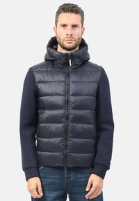 Gilet imbottito blu navy con cappuccio, chiusura con zip e maniche a coste blu navy, con una texture liscia e un motivo a righe orizzontali.