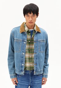 Denim jas in lichtblauw met een corduroy kraag, knoopsluiting aan de voorkant en twee borstzakken. Gelaagd over een groen geruit overhemd.