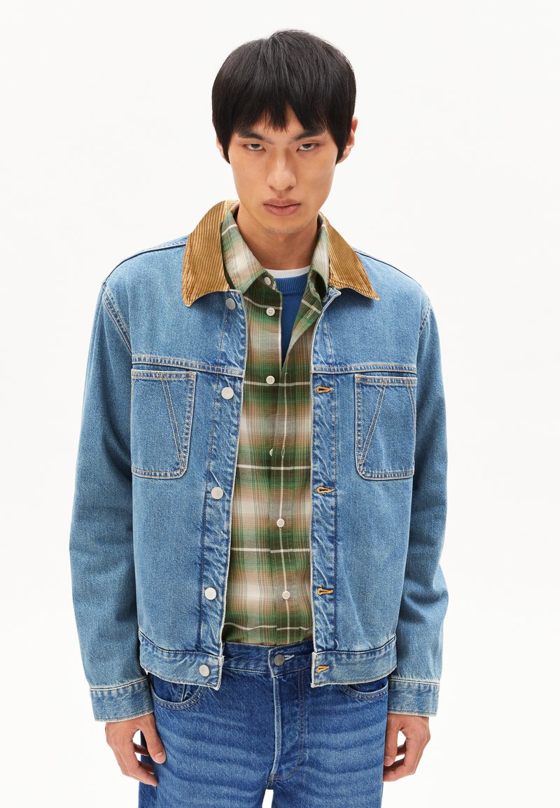 Denim jas in lichtblauw met een corduroy kraag, knoopsluiting aan de voorkant en twee borstzakken. Gelaagd over een groen geruit overhemd.