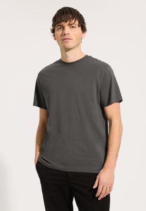 Een jonge man staat met zijn handen in zijn zakken, draagend een donkergrijs T-shirt met korte mouwen en een zwarte broek, tegen een effen achtergrond.