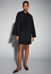 Veste noire courte portée sur un haut noir ajusté, associée à une jupe noire longueur genoux et des mules à talons noires, sur un fond neutre.