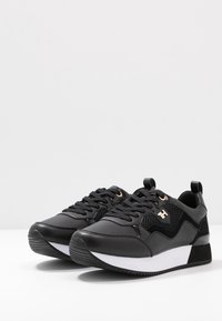 Chaussures de sport noires en cuir synthétique et mesh, avec une semelle intermédiaire blanche, une surface texturée et des accents dorés sur les lacets.