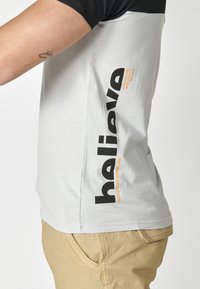 Siva kratka majica s crnim detaljima, s velikim crnim natpisom sa strane i narančastim tekstualnim detaljem. Mekani materijal, opušteni kroj.