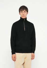 Pull noir à col zippé avec un col et des poignets côtelés. Fabriqué en matériau doux, il présente une coupe décontractée et une couleur unie distinctive.