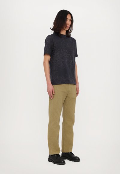 Tricou negru cu mânecă scurtă, cu un model texturat, asociat cu pantaloni din cordură khaki și pantofi negri cu șireturi. Fundal simplu.