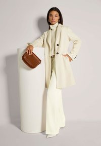 Vrouw in een crèmekleurige jas, turtleneck, wijde broek en witte hakken leunt op een witte cilindrische sokkel, terwijl ze een bruine handtas vasthoudt met een paar grote oorbellen.