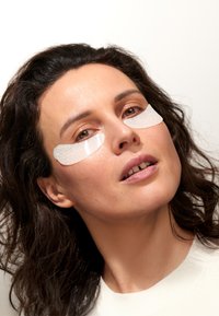 Hydrożelowe płatki pod oczy w przezroczystym, błyszczącym materiale nałożone pod oczami. Ciemne, falujące włosy otaczające zbliżenie na twarz na jasnym tle.