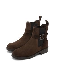 Botas de tobillo de ante marrón con una suela de goma gruesa, paneles laterales elásticos y un detalle de hebilla en la correa que cruza el tobillo.