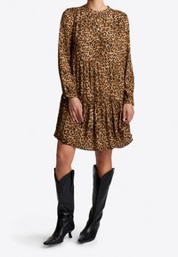 Vestido de estampado de leopardo con mangas largas y diseño de falda en capas. La tela parece ligera. Botas de cowboy negras hasta la rodilla con punteras puntiagudas.