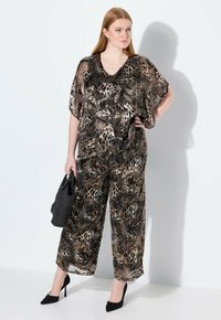 Vrouw in een bijpassende losvallende blouse en wijde broek van luipaardprint, met een zwarte handtas, staande tegen een witte muur.