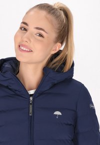 Marineblauwe gewatteerde jas met capuchon, met een zilveren paraplu-logo op de borst. Vlotte textuur, volledige rits en doorgestikte ontwerp.