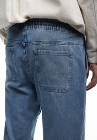 Jeans de mezclilla azul claro con una cinturilla elástica, que presentan dos bolsillos traseros y un corte relajado. La tela tiene un acabado suave y ligeramente texturizado.