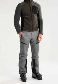 Veste en polaire avec des panneaux gris et verts, accents noirs, zip intégral et poche poitrine ; associée à un pantalon gris avec poches zippées et côtés noirs.