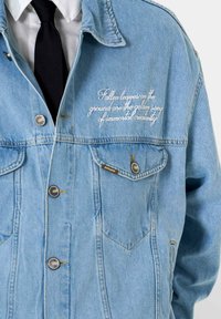 Veste en denim bleu clair avec deux poches poitrine, boutons argentés et texte brodé en cursive blanche sur le côté gauche.