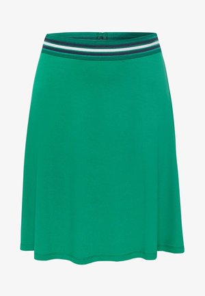 Vrouw die een groene rok draagt, wit mouwloos topje, gedeeltelijk uitgetrokken blauwe spijkerjasje en witte sneakers, staand met gespreide benen tegen een witte achtergrond.