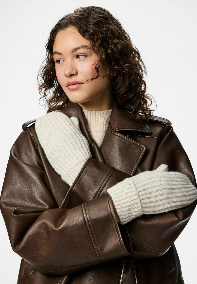 Cremefarvede ribbede vanter lavet af blødt strikkemateriale, båret over en brun læderjakke med strukturerede skuldre og klapdetaljer.