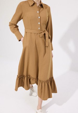 Brązowa sukienka midi z długim rękawem, zapinana na guziki z przodu, z paskiem w talii i falbaną na dole, noszona z białymi sznurowanymi butami na jasnym tle.
