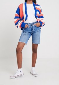 Färgglad randig kofta i orange, blått och vitt; vit t-shirt; högmidjade denimshorts; vita sneakers med stjärnlogo.