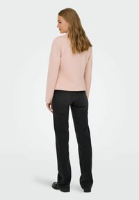 Femme aux cheveux longs portant un blazer près du corps de couleur rose clair, un jean noir à jambes droites et des chaussures noires, debout et de dos.