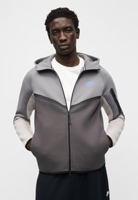 Pelēks sporta džemperis ar rāvējslēdzēju un kontrastējošām daļām, ar kapuci, melniem akcentiem un zilu Nike logotipu uz krūtīm. Kombinēts ar melnām biksēm.