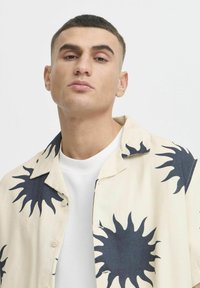 Camicia button-up crema con motivi solari blu scuro, dal design collo casual. Indossata sopra una camicia bianca con texture.