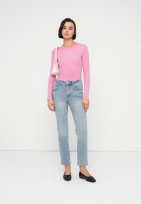 Ροζ μακρυμάνικο τοπ, ανοιχτό μπλε τζιν με ίσια ποδιά, μαύρες μπαλαρίνες. Το μοντέλο κρατά μια μικρή λευκή τσάντα χειρός, μινιμαλιστικό στυλ, μπροστά σε απλό φόντο.