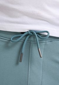 Lichtgroene sweatpants met een trekkoord in de taille. Het trekkoord heeft metallic-accenten aan de uiteinden. De stof is soepel met subtiele stiksels.