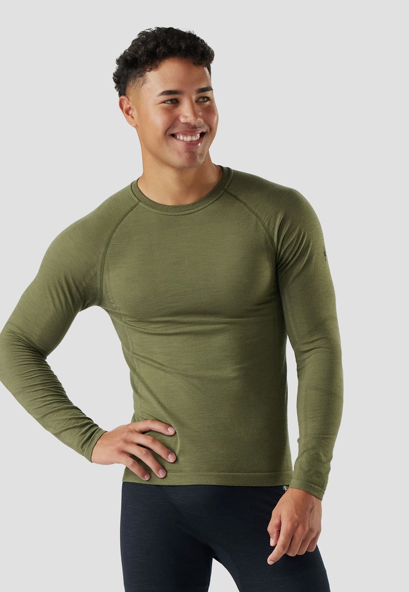 Mladý muž se usmívá, má na sobě olivově zelené přiléhavé tričko s dlouhým rukávem a tmavé sportovní kalhoty, stojí s jednou rukou v bok.