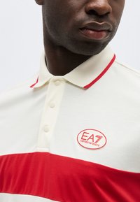 Κοντινό πλάνο ενός ατόμου που φοράει λευκό πόλο πουκάμισο με κόκκινη οριζόντια ρίγα και λογότυπο EA7 Emporio Armani στο στήθος.