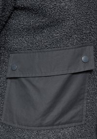 Tissu polaire texturé noir avec une poche plaquée gris foncé dotée de deux boutons-pression, accentuée par une surface lisse.