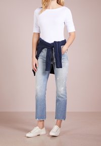 Bílé přiléhavé triko s krátkým rukávem v kombinaci s vybledlými světle modrými džíny se třepenými okraji. Tmavě modrý pletený svetr uvázaný kolem pasu. Bílé tenisky.