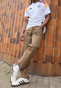 Person som bär en vit tysk fotbollsjacka med färgglada detaljer, bruna vida byxor och vita Adidas-sneakers, stående mot en kaklad vägg.