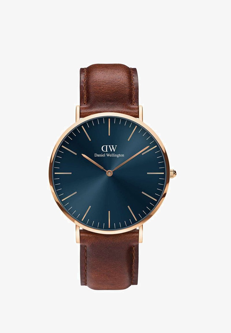 Tume sinise kelladialiga pruun nahkrihmaga kell, millel on kuldse tooniga tunnimarkerid ja näpitsad. Omab minimalistlikku disaini sujuvate metallaktsentidega.