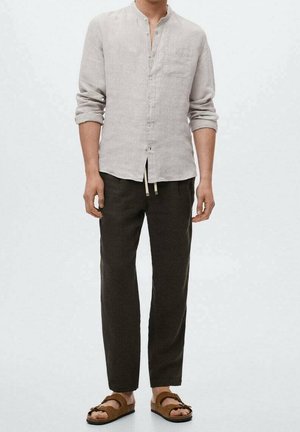 Man draagt een beige, knoopjes lang mouwen overhemd, donkere broek en bruine sandaalopen tenen tegen een effen lichte achtergrond.