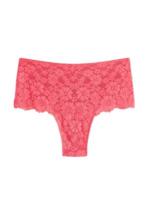 Kvinde med langt krøllet hår står barfodet iført koralrosa blonder-bralette og højttaljede trusser foran en ensfarvet lys baggrund.