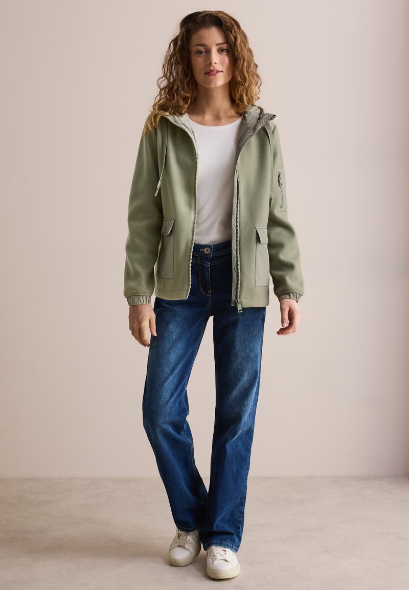 Giacca a zip verde chiaro con cappuccio, due tasche frontali e fodera grigia, indossata sopra una camicia bianca e jeans blu scuro. Sneakers bianche.