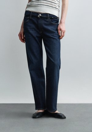 Personne portant un jean droit bleu foncé avec une ceinture noire, des chaussures plates noires et un haut tricoté sans manches blanc, debout sur un sol gris.