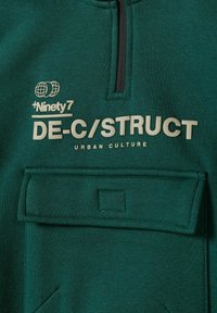 Sötétzöld pulóver félig cipzáros kivitelben, elülső zsebből és bézs nyomtatott szövegből áll: "DE-C/STRUCT URBAN CULTURE" és logók.