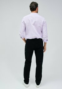 Camicia di lino viola chiaro a maniche lunghe con polsini arrotolati; abbinata a pantaloni neri aderenti con tasche posteriori e un accento blu.
