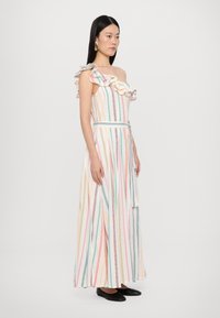 Vestido maxi com babados à mostra nos ombros, apresentando riscas verticais em tons pastel, uma cintura atada e um tecido leve. Sapatilhas pretas usadas por baixo.