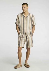 Πουκάμισο με κουμπιά και ρίγες σε μπεζ, καφέ και κρεμ αποχρώσεις· σορτς που ταιριάζουν. Φορεμένα με μπεζ σανδάλια. Κοντά μανίκια, χαλαρή εφαρμογή και τσέπες.