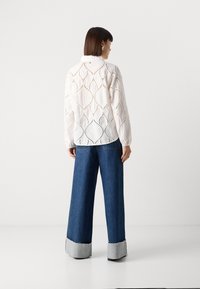 Blouse blanche avec découpes géométriques et manches longues, associée à un jean en denim bleu taille haute et large avec des poignets gris contrastants.