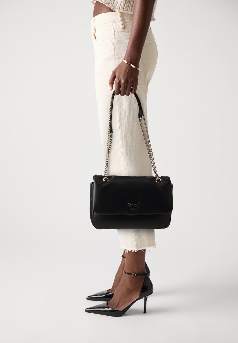 Donna in jeans bianchi e tacchi neri tiene una borsa a tracolla nera con catena e hardware dorato su uno sfondo bianco.